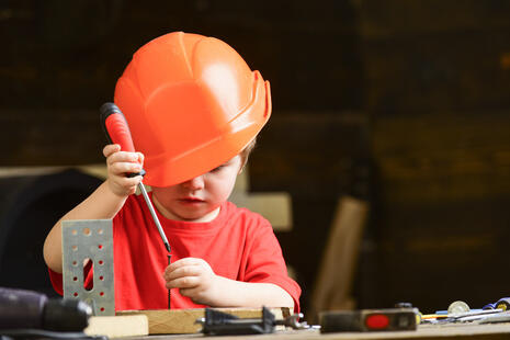 Ein Kind mit einem organgen Bauhelm hantiert mit einem Schraubendreher. Vor dem Kind liegen Werkzeuge.